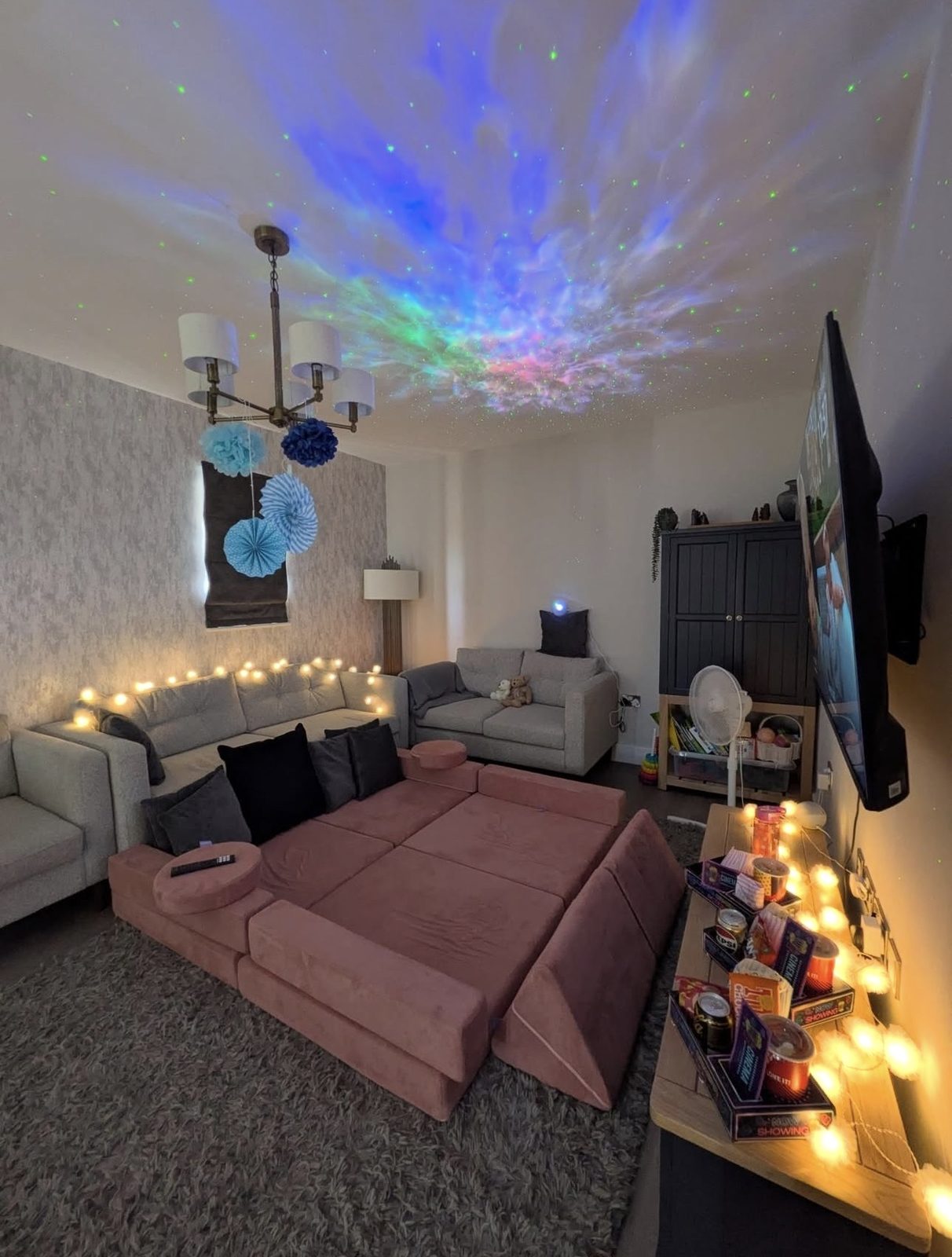 Cosy living room with star projector on the ceiling, fairy lights and neon snack trays by the sofa