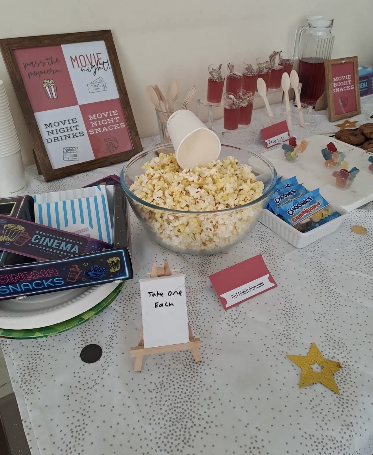 Party table with a bowl of popcorn, movie night sign, drinks and neon snack trays at the edge