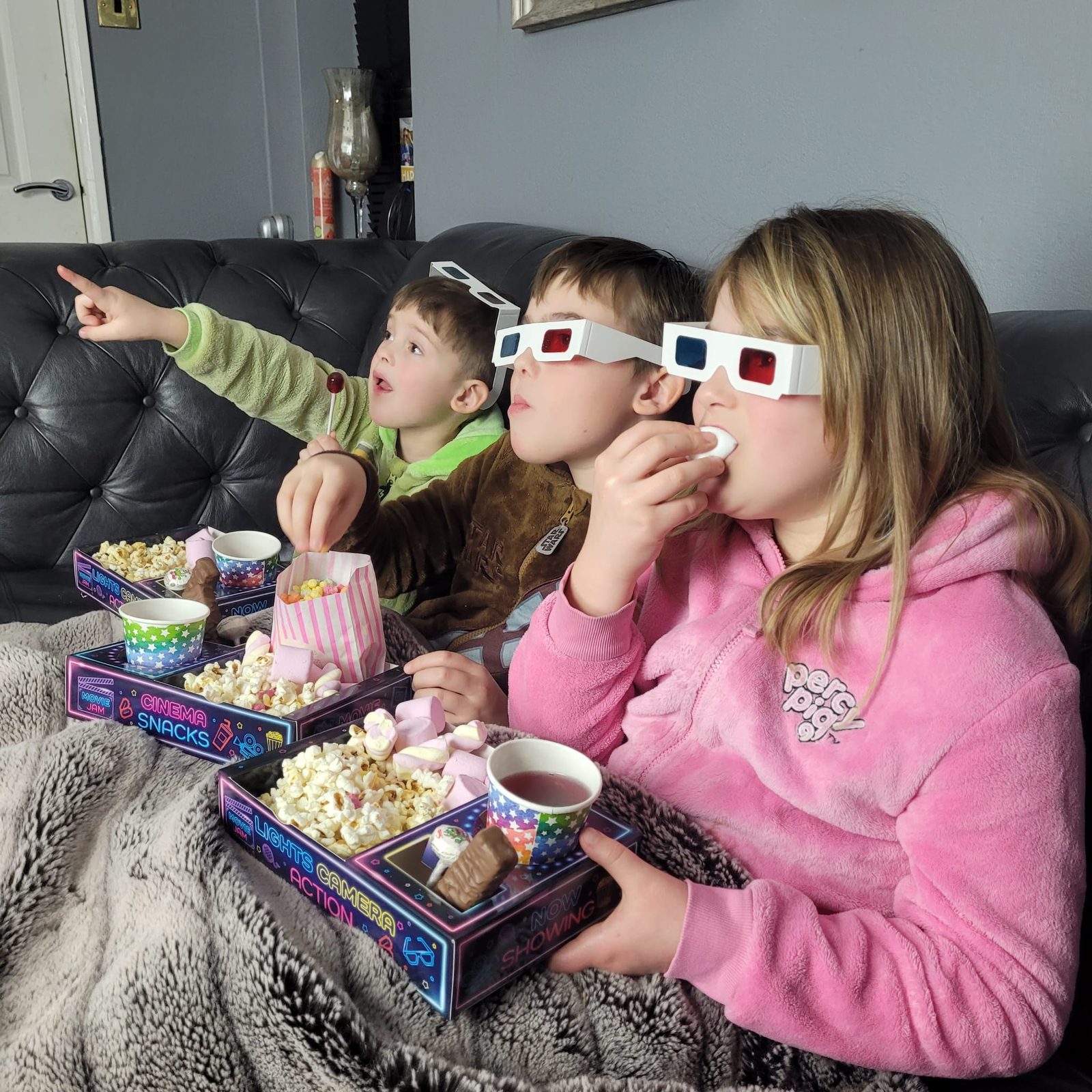 Three children in 3D glasses under a blanket on a leather sofa eating from snack trays