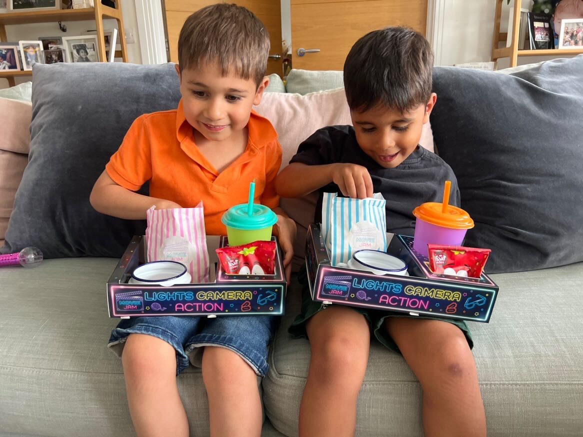Two smiling boys on a grey sofa each holding a neon snack tray with popcorn and cups
