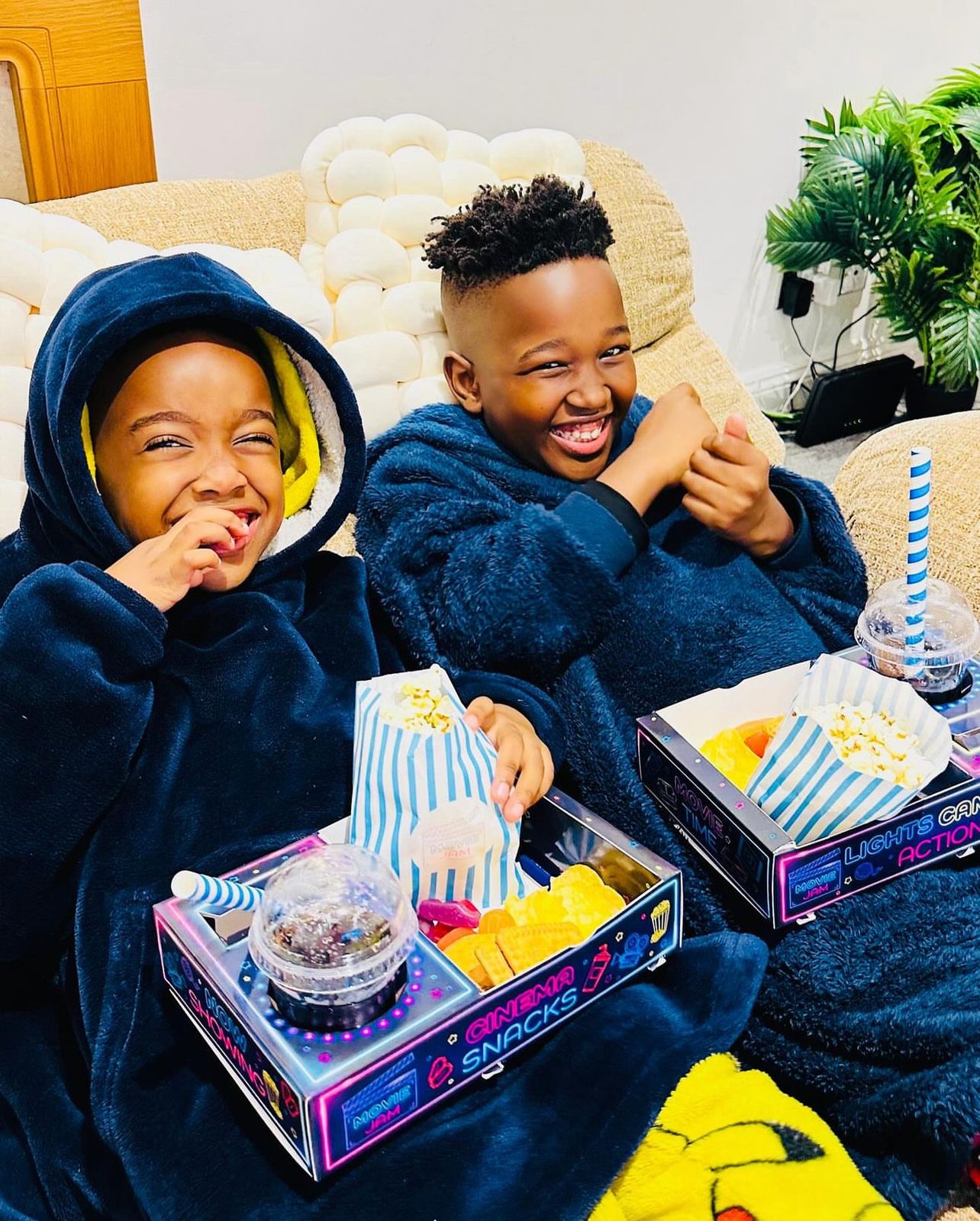 Two laughing children in dark hooded blankets on a sofa with striped popcorn bags and a snack tray