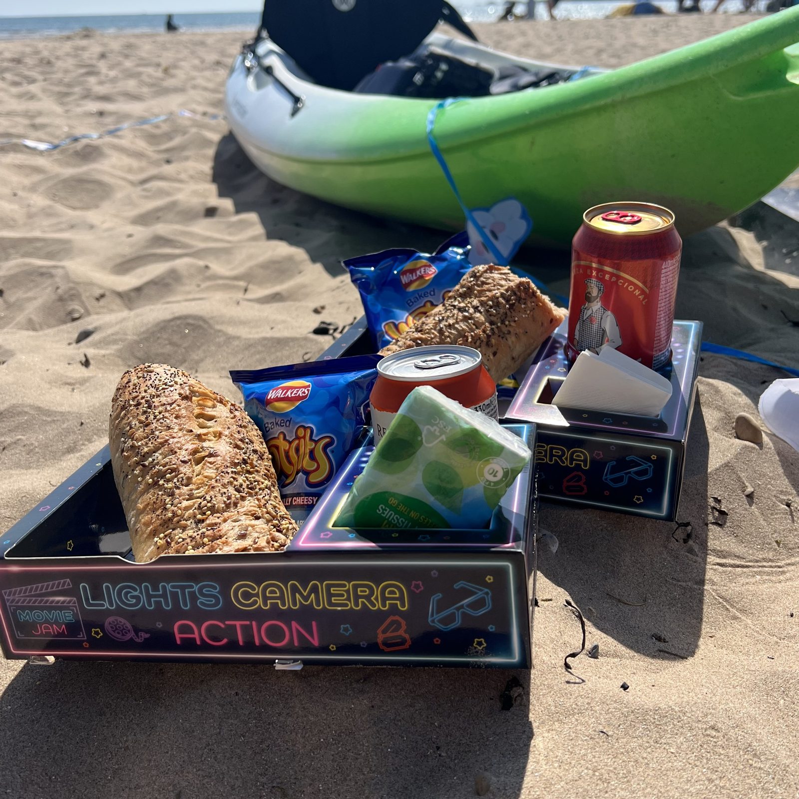 Snack trays on a sandy beach beside a green kayak, holding bread, crisps and a drink can