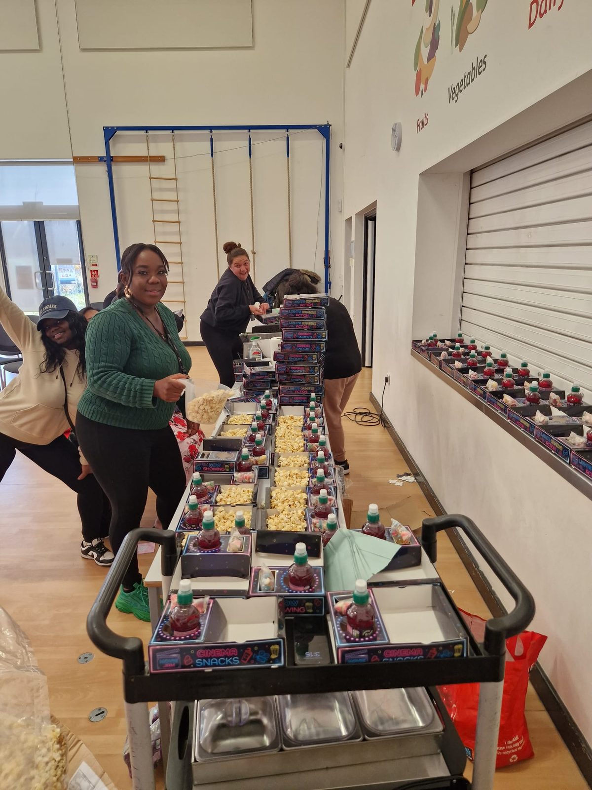 Adults preparing rows of filled snack trays on trolleys and tables in a school-style hall