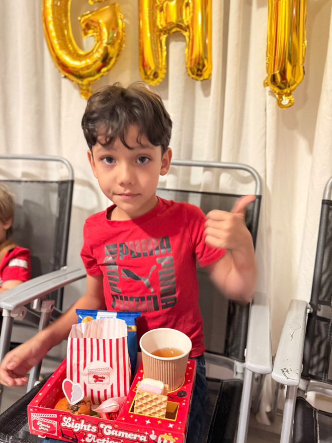 Smiling boy in red shirt giving thumbs up, holding a red snack tray with popcorn and a drink