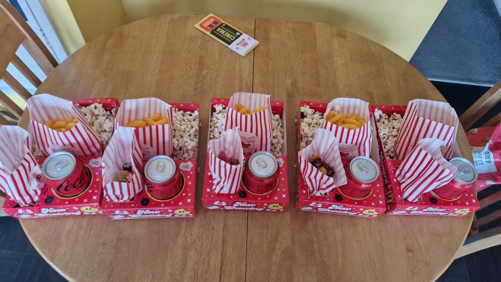 Five red snack trays with popcorn, crisps and cans lined up on a round wooden table