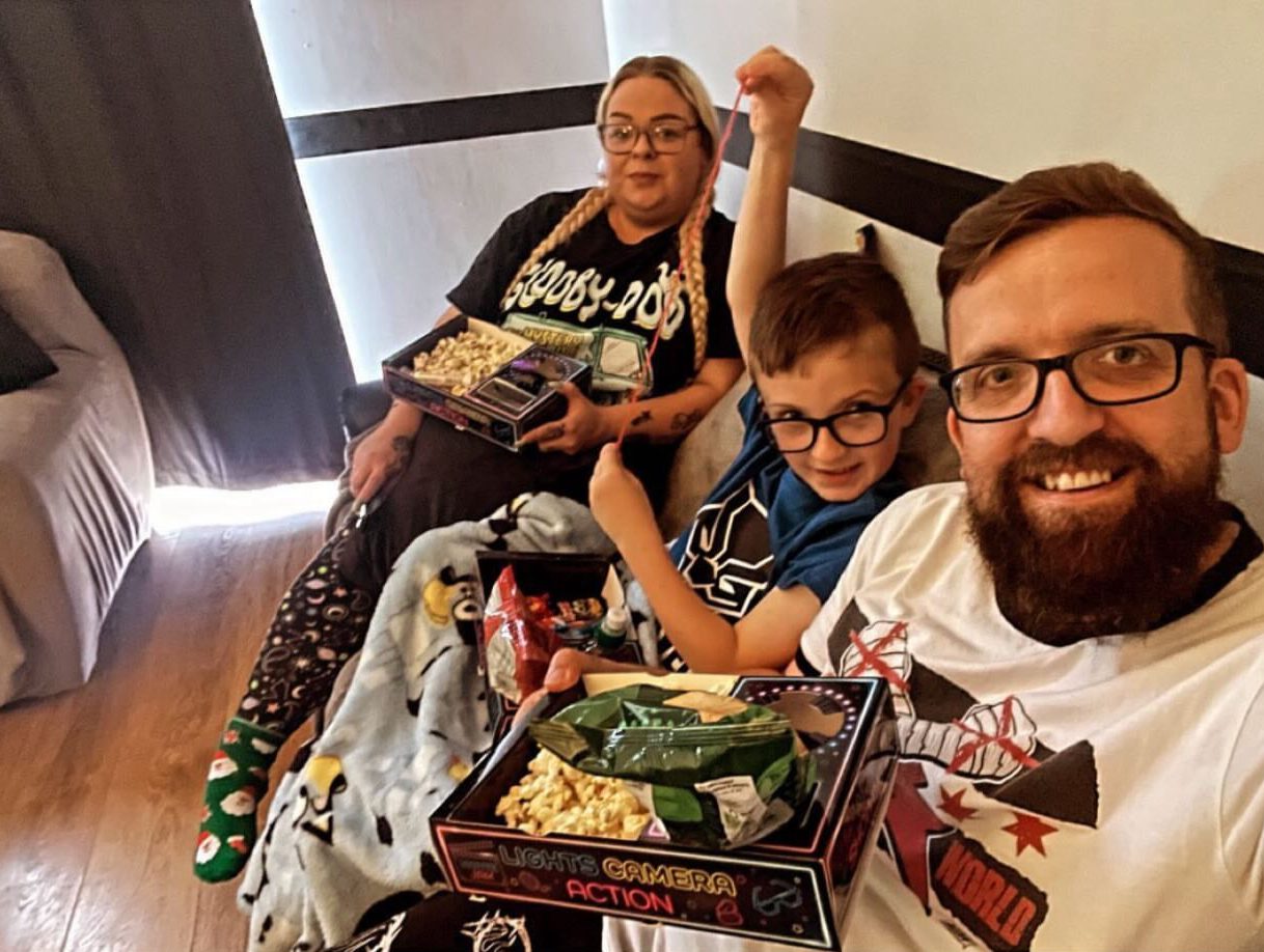 Family of adults and a child on a sofa with blankets sharing a snack tray in front of the TV