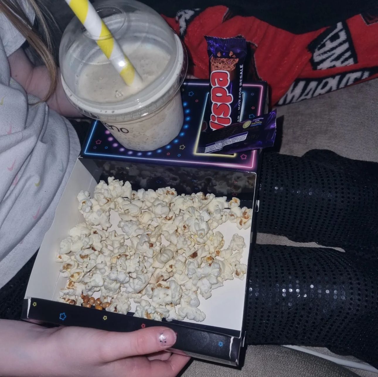 Hands holding a snack tray of popcorn beside a milkshake with a straw, on a lap
