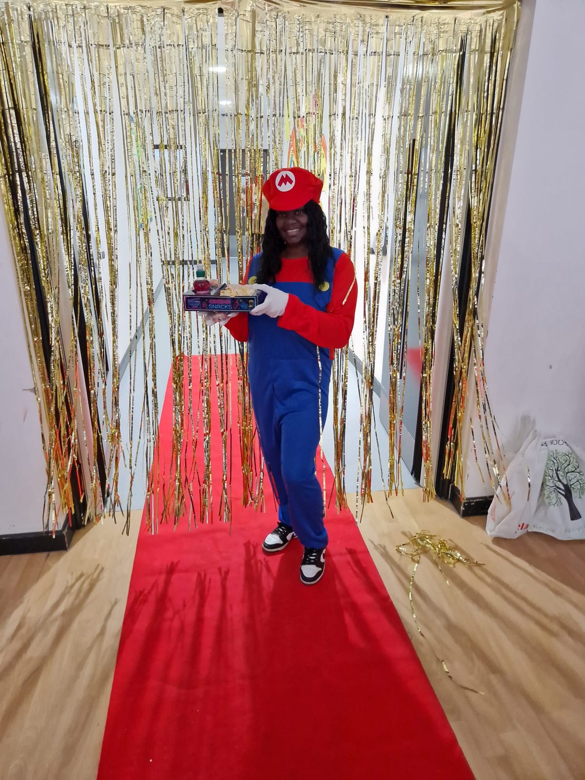 Person in a red Mario-style costume carrying a snack tray along a red carpet with foil curtain