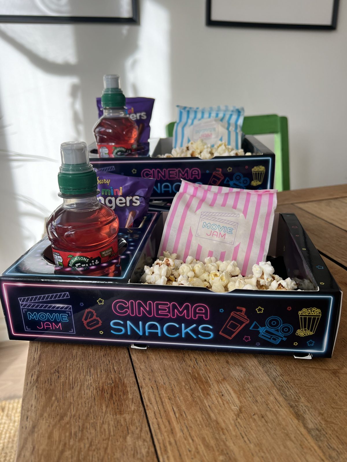 Neon Cinema Snacks tray on a wooden table with popcorn, striped bags and a drink bottle