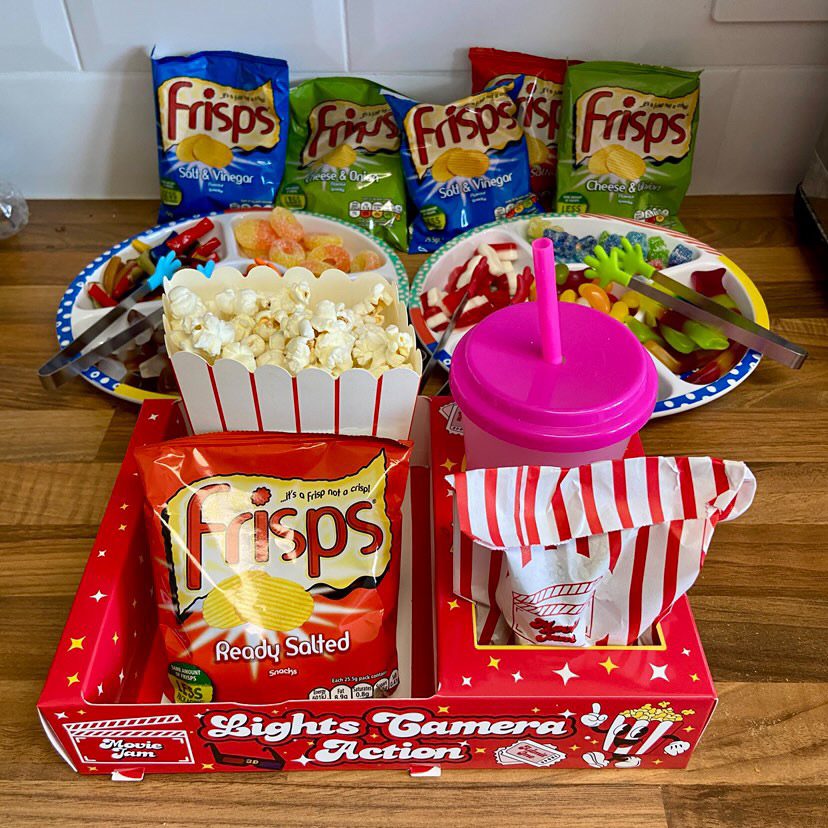 Red snack tray with popcorn, crisps and a pink cup on a table with more sweets behind